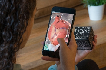 Over the shoulder view of a girl holding a smartphone in one hand and MERGE augmented reality cube in another. The smartphone displays a holographic heart.