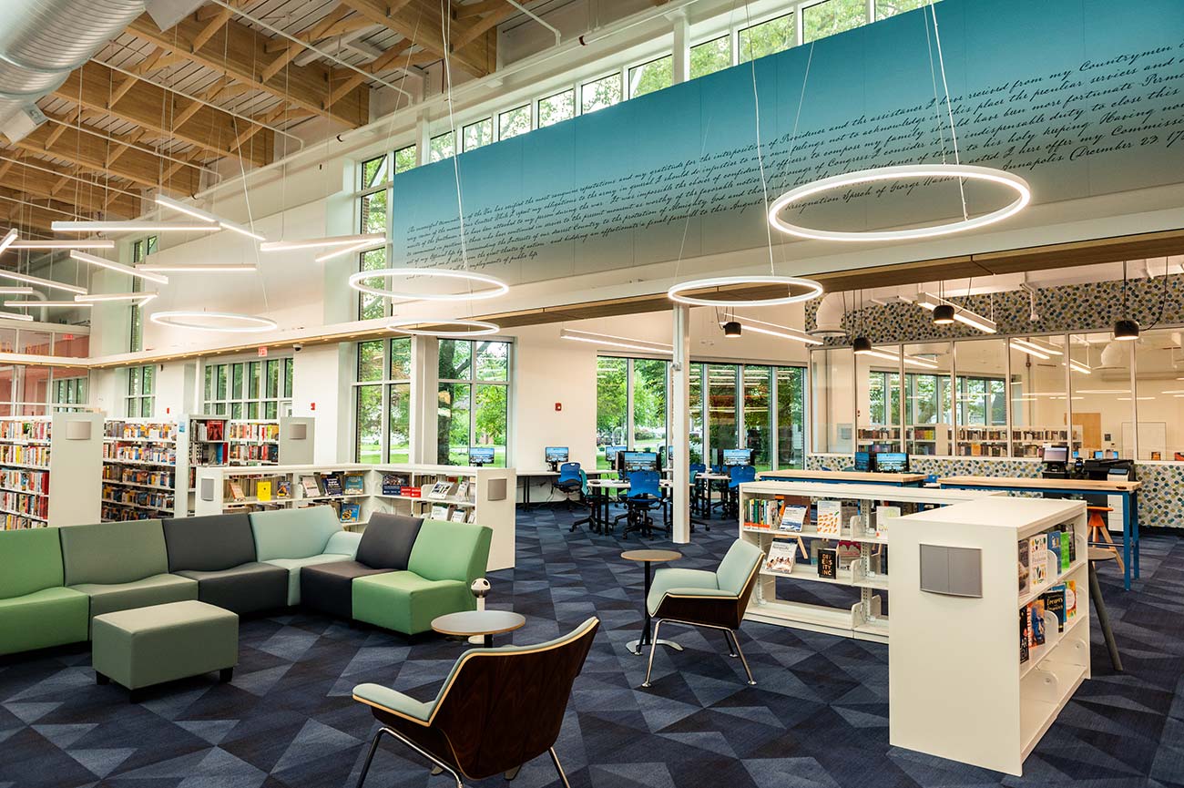 interior of Busch Annapolis Library with seating, shelves holding books, circular lighting, geometric carpet, exposed beams at ceiling, windows along wall