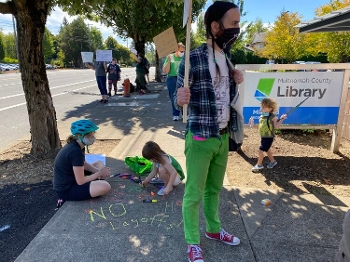 man in mask holding protest sign in front of Multnomah County Library sign with protesters in background, child and masked parent on sidewalk writing