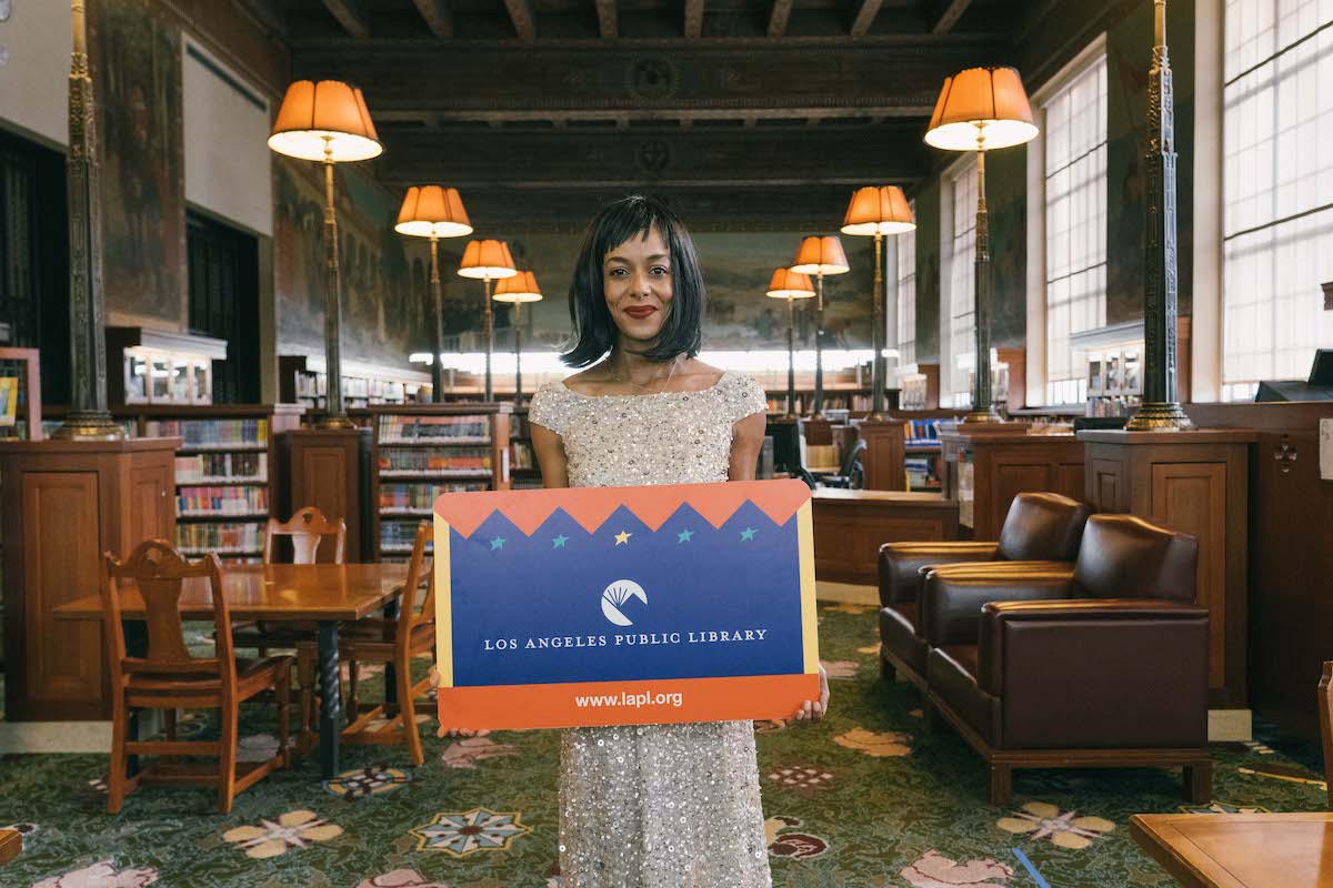 Lisa Lucas in glittery evening gown standing in library with bookshelves on either side of aisle, holding sign that says