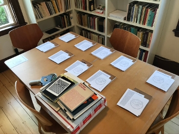 Piles of paper material and books arrayed on desk, more books stacked on mailing box with tape gun alongside
