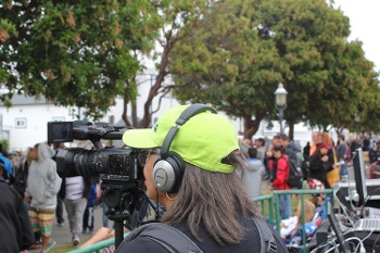 woman wearing baseball cap seen from behind, filming crowd with handheld camera