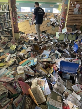 man stepping over room littered with books covering the floor, fallen bookcases