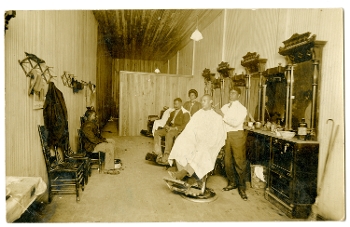 sepia tone postcard showing African American barbershop, two barbers (man and woman) behind chairs and young man sitting against wall