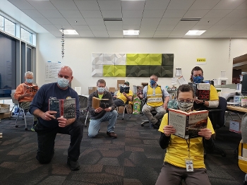 7 men wearing masks, seated or kneeling in library room, reading books