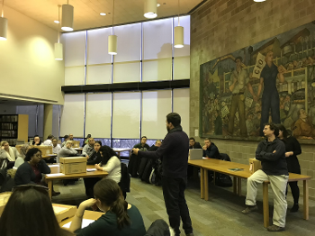 students sitting in chairs and on desks watching man in center lecturing in library with labor-themed mural on one wall