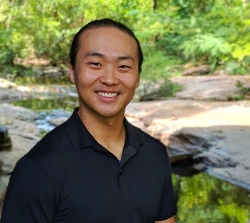 DeLa Dos head shot - Man in black shirt smiling against green outdoor background