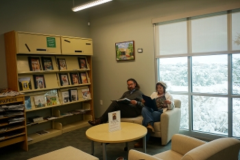man and woman, seated, reading periodicals in library with snowy vista outside large windows